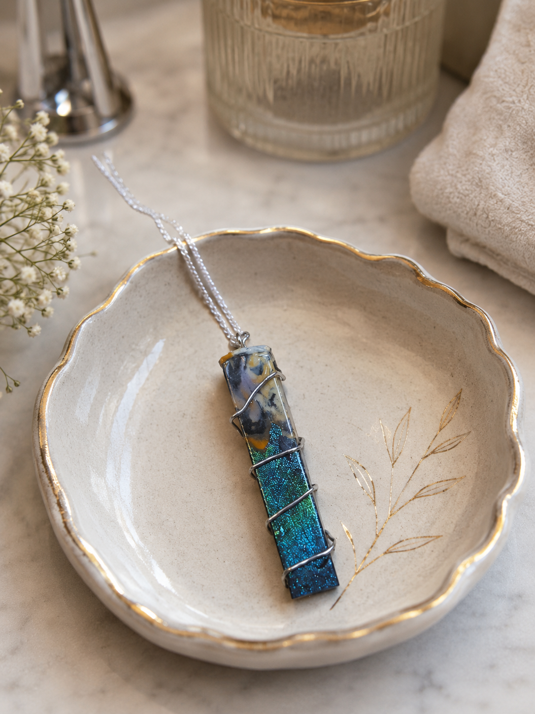 A rectangular pendant necklace with a blue and teal marbled stone wrapped in silver wire, resting on a scalloped cream ceramic dish with gold detailing.