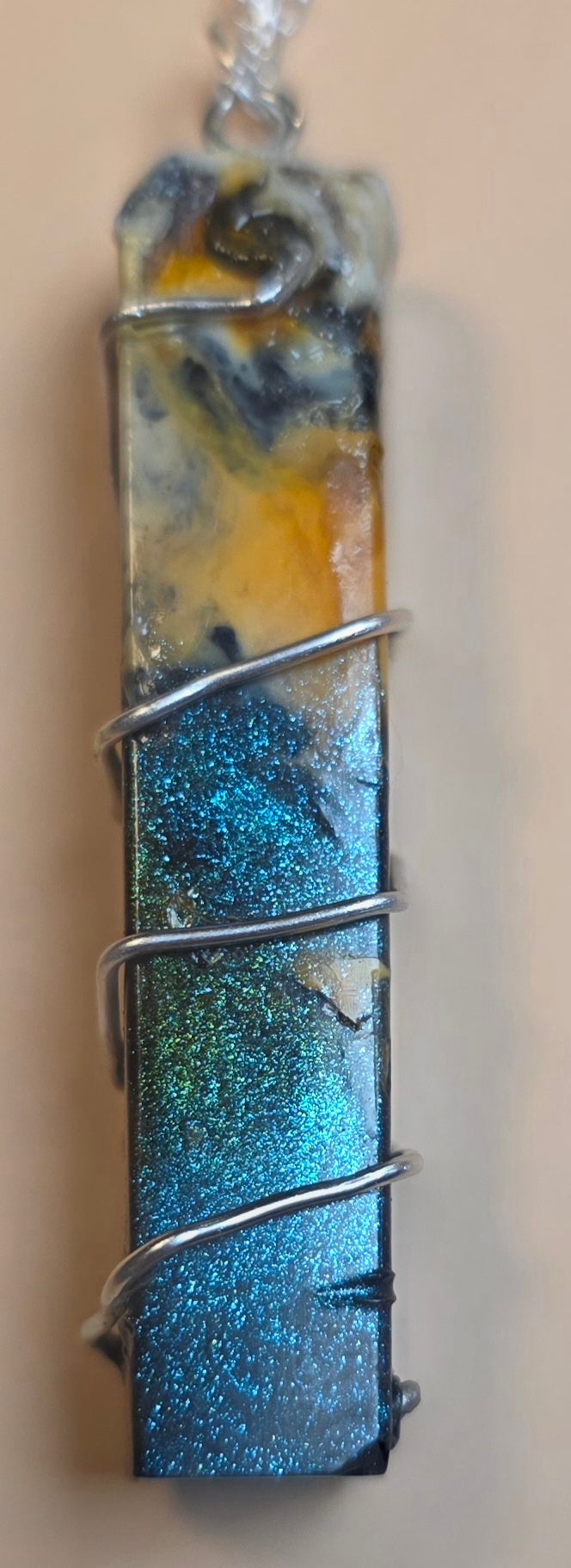 A rectangular resin pendant necklace wrapped in silver wire, featuring a marbled design with blue, yellow, and black, and a glittery blue lower section, shown against a plain beige background.