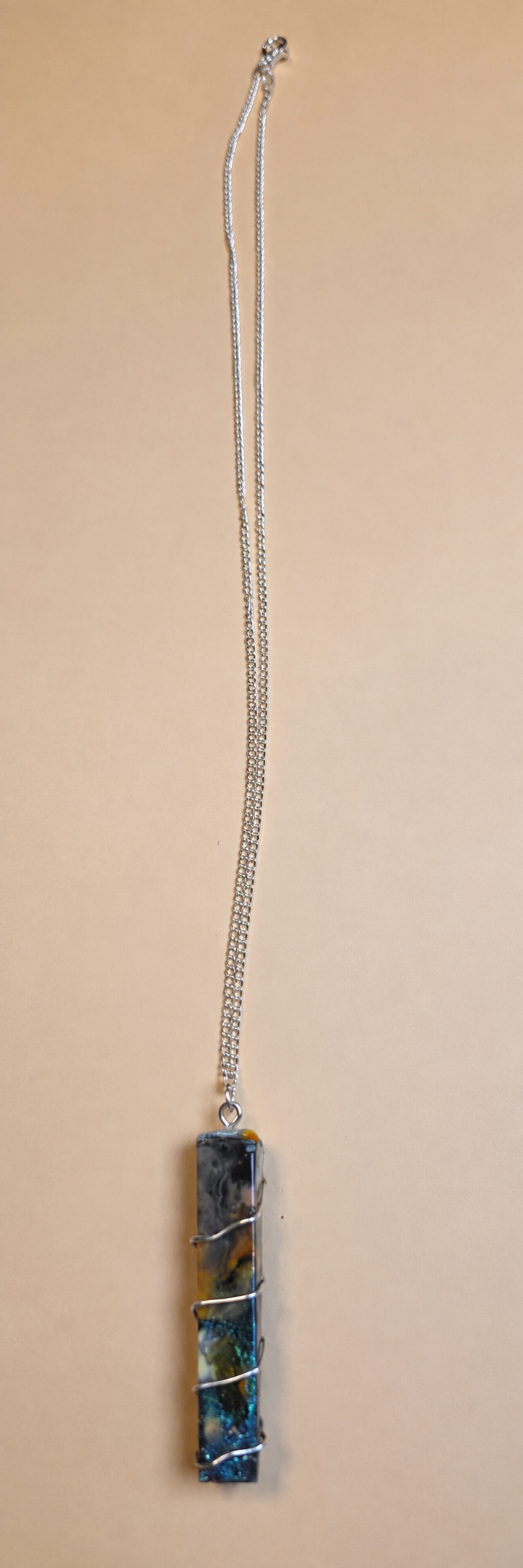 A silver chain necklace with a vertically oriented rectangular pendant wrapped in silver wire, featuring a blue and green marbled stone, shown against a solid beige background.