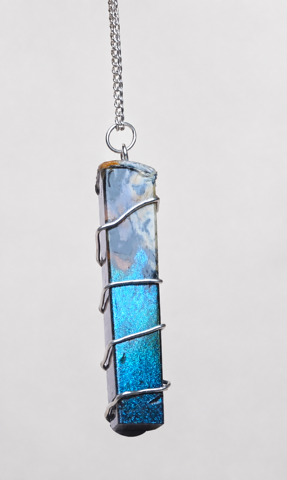 A vertical rectangular pendant necklace featuring a glittery blue and white stone wrapped in silver wire, suspended from a silver chain against a light gray background.