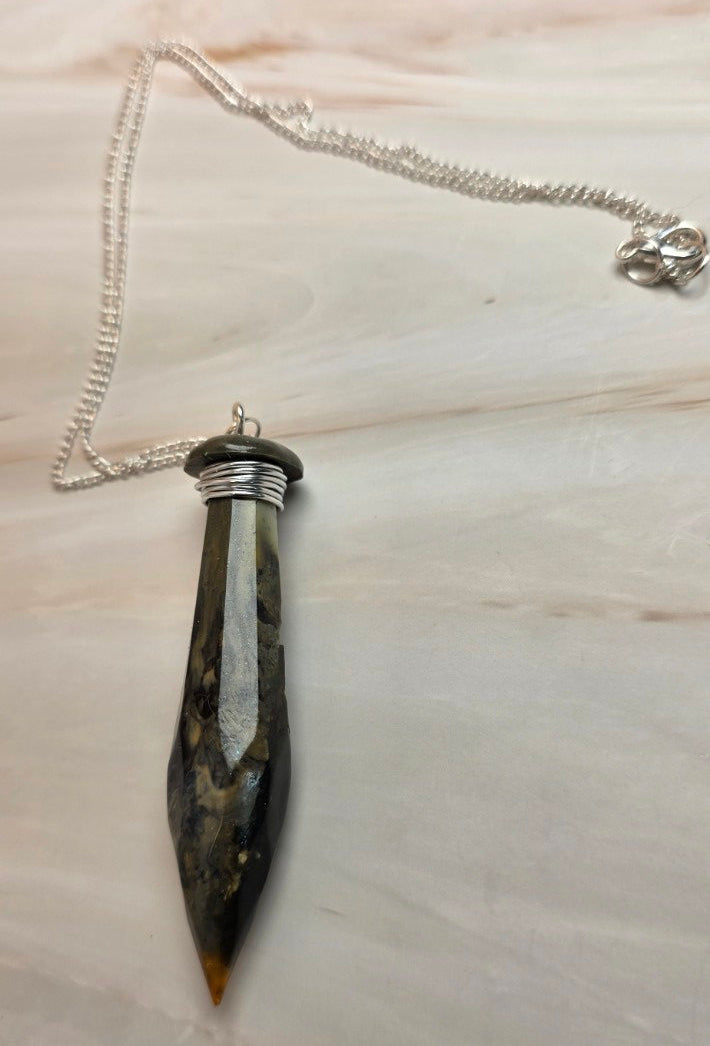 A pointed dagger pendant made of dark brown and black wood with a silver blade, attached to a silver chain, displayed on a light wood background.
