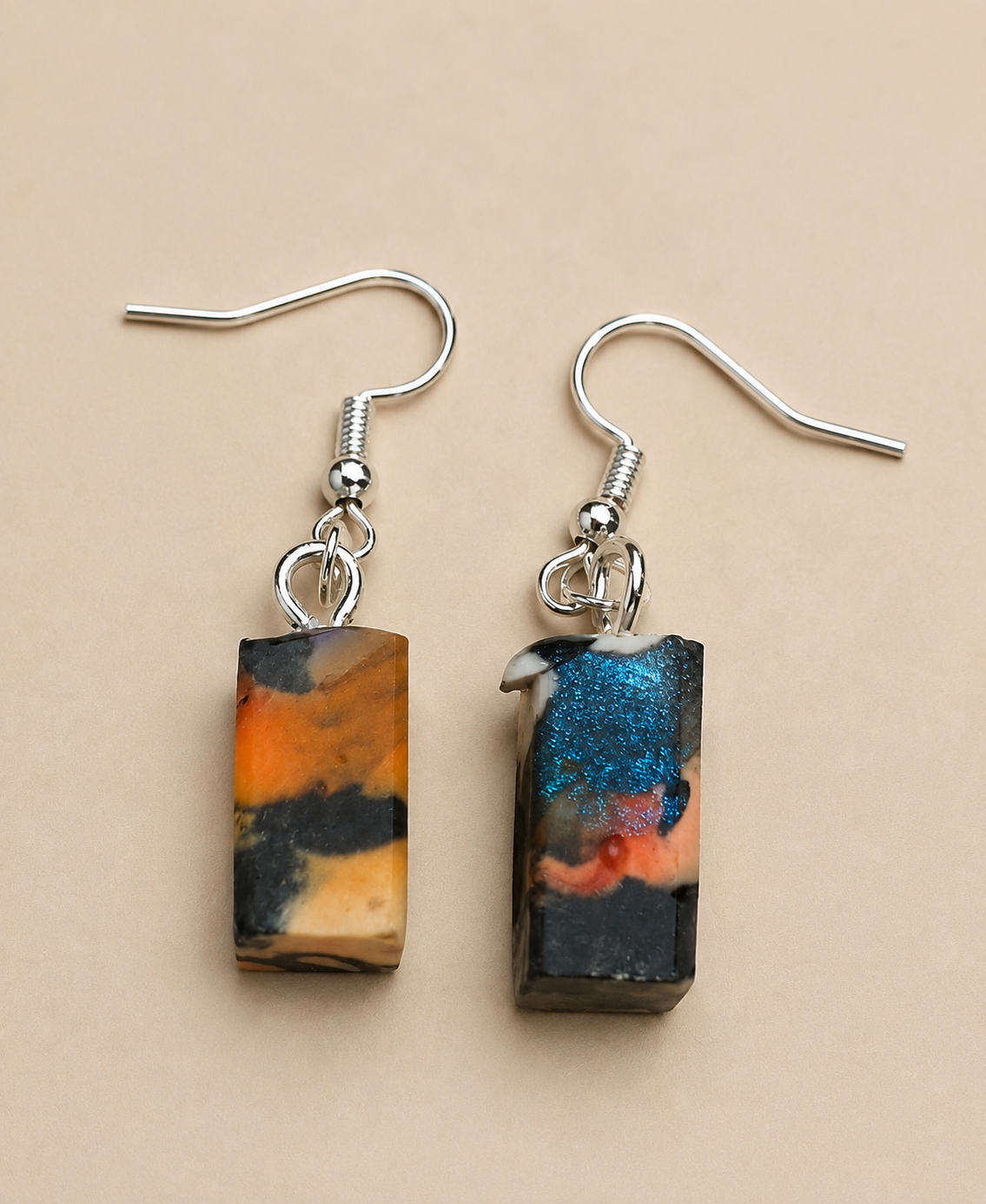 A pair of rectangular drop earrings with silver fishhook wires, featuring marbled resin pendants in orange, black, beige, and a glittery blue section, displayed on a solid beige background.