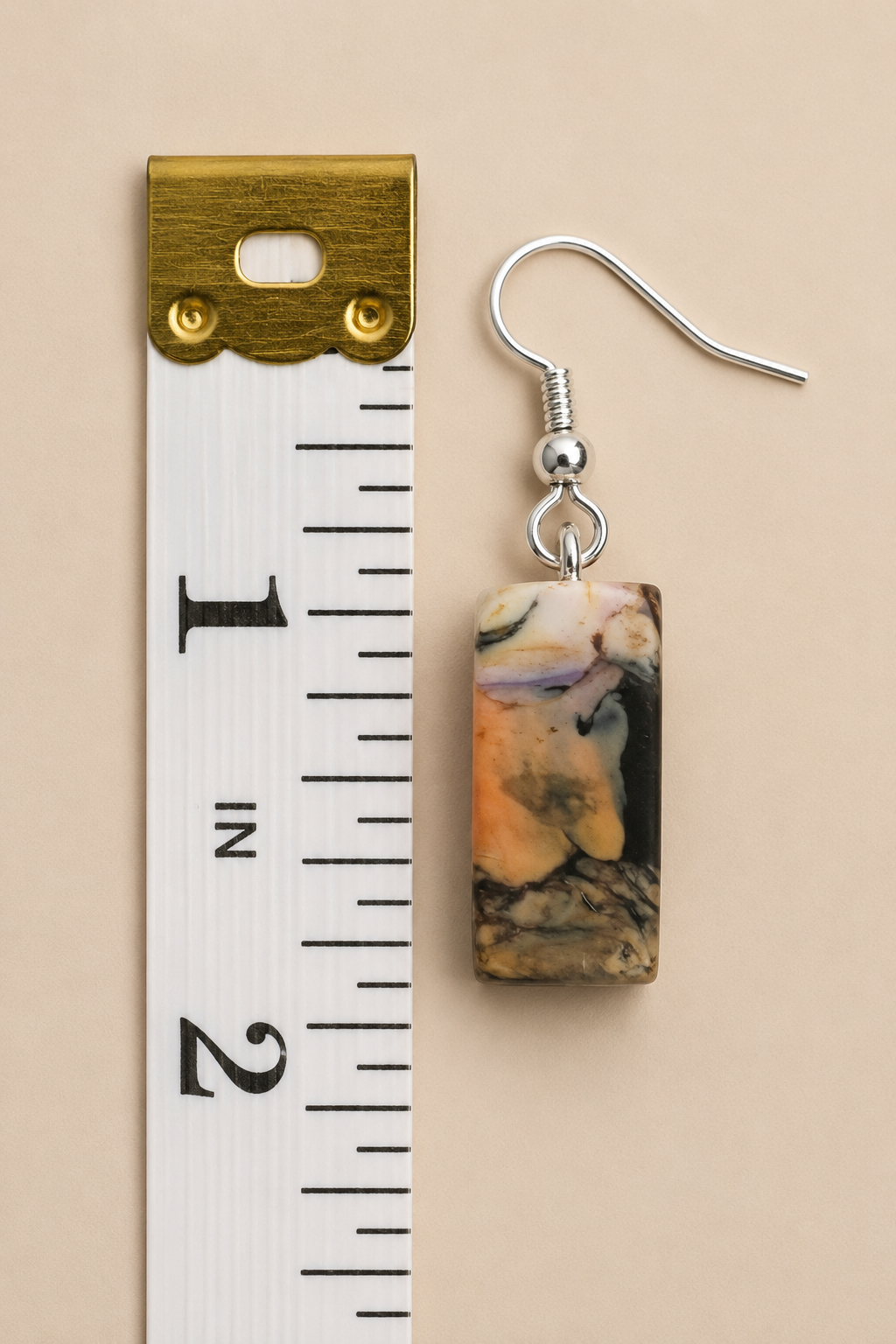 A single rectangular drop earring with a marbled stone-like pattern in beige, brown, black, and soft lavender, hanging beside a white measuring tape on a warm beige background.