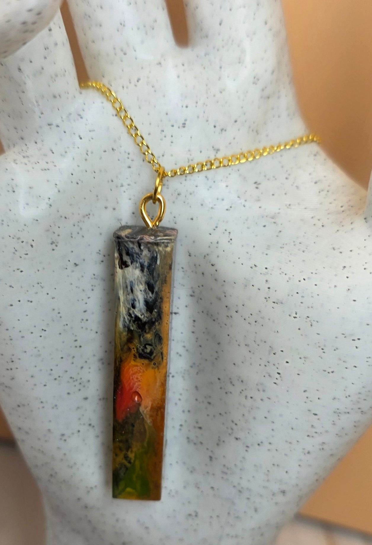 A rectangular resin pendant necklace with a colorful landscape design, hanging from a gold chain on a white speckled hand-shaped jewelry display.