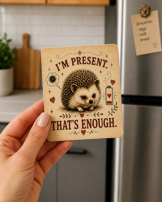 A hand holds a square refrigerator magnet in a kitchen setting. The magnet features a vintage-style illustration of a hedgehog with the text 'I'M PRESENT.