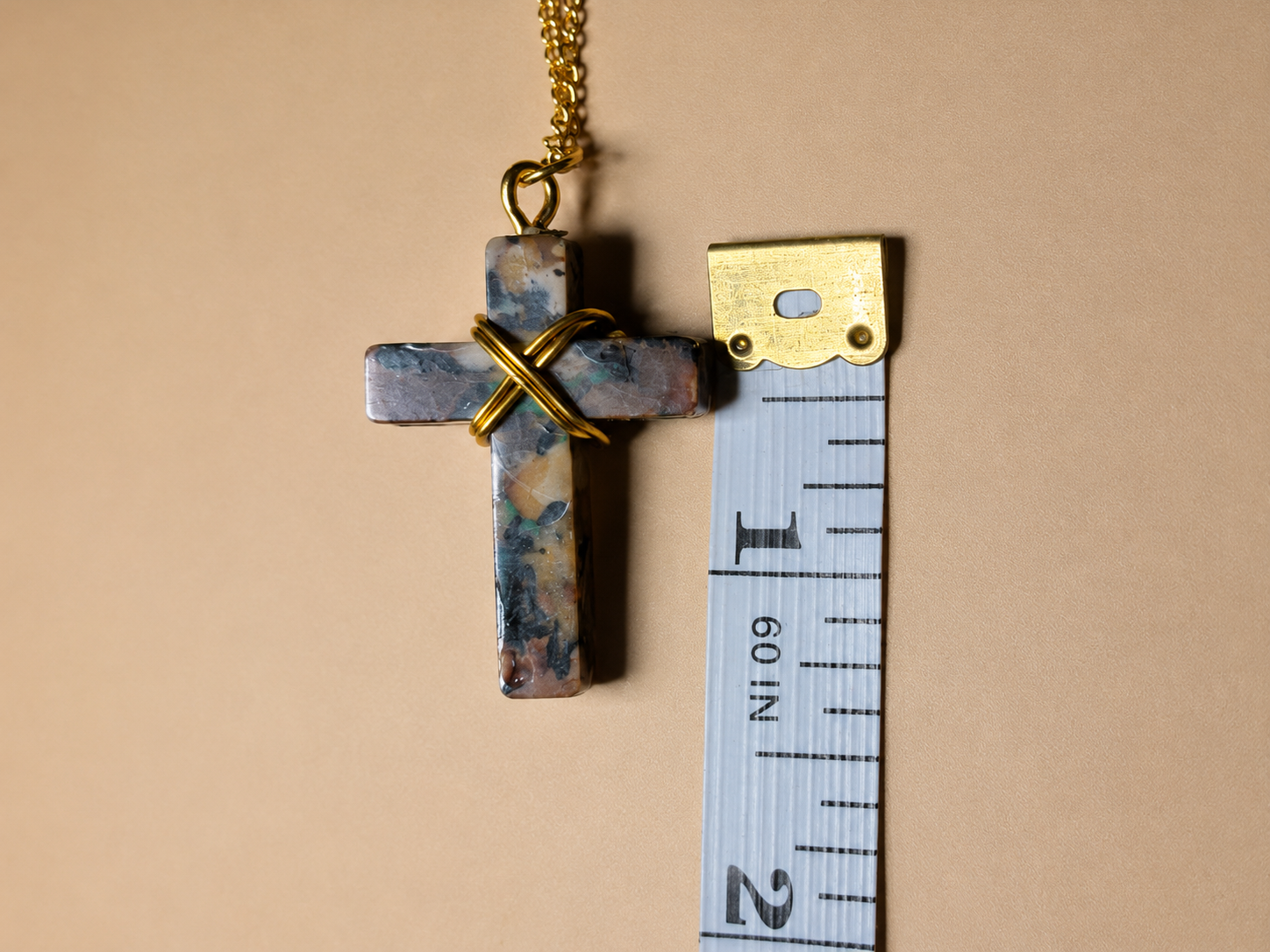 A cross-shaped pendant necklace made of marbled blue, gray, and white stone with a gold wire cross wrapped over the center and a gold chain, shown next to a measuring tape on a beige background.