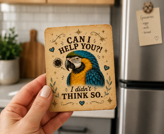 A hand holds a square decorative coaster featuring a colorful parrot illustration and the text 'CAN I HELP YOU?! I didn't THINK SO.' in a kitchen setting with a refrigerator and countertop blurred in the background.