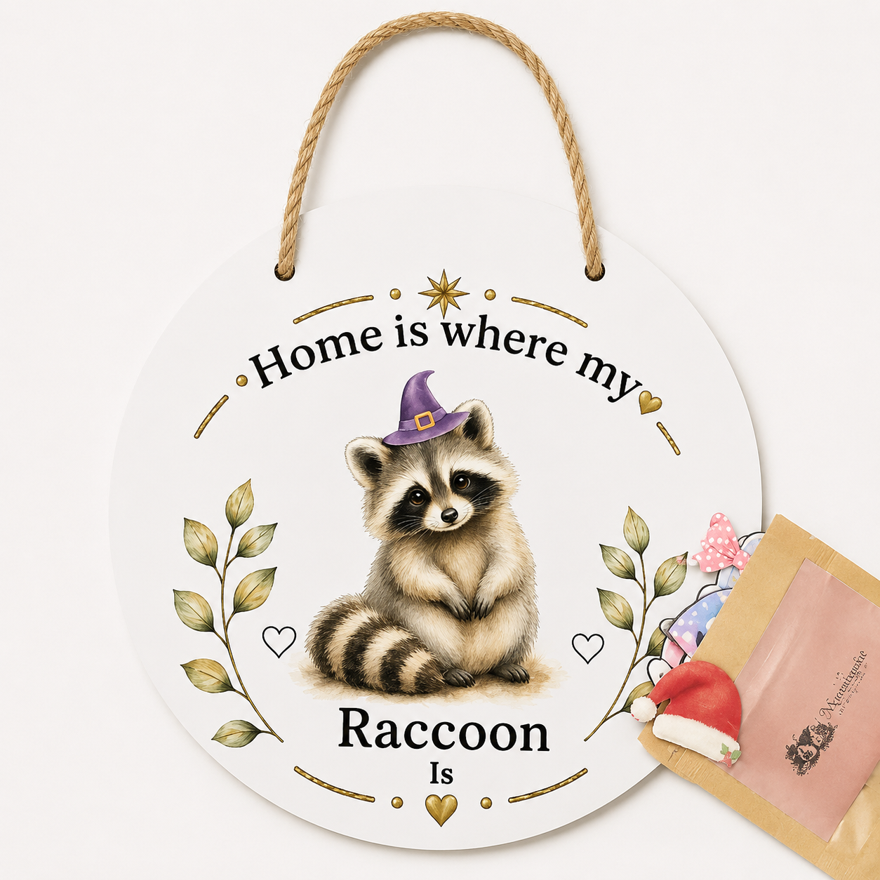 A round white hanging plaque with a rope handle, featuring an illustrated raccoon wearing a purple witch hat and the text 'Home is where my Raccoon Is.' A small gift bag with a Santa hat and pink ribbon sits beside it on a clean white background.