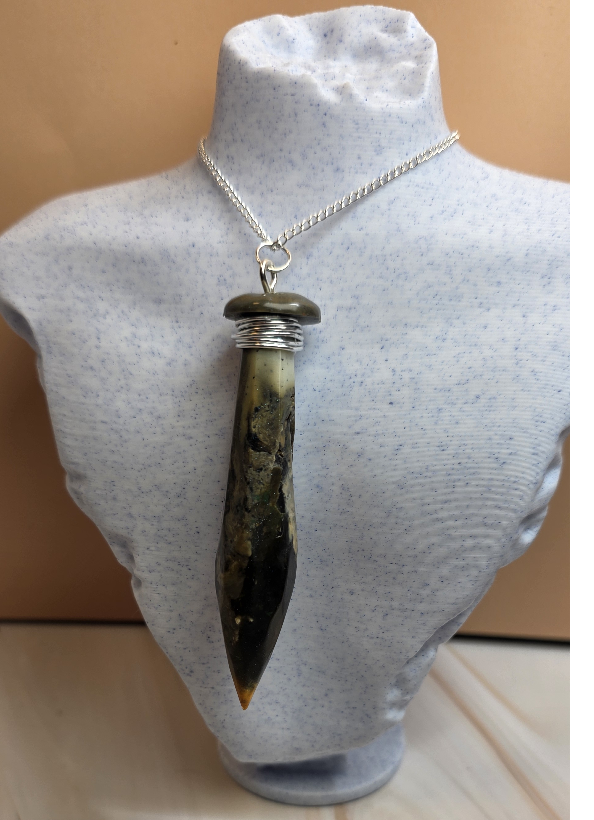 A silver chain necklace with a pointed polished stone pendant, displayed on a light blue speckled jewelry bust against a warm beige background.