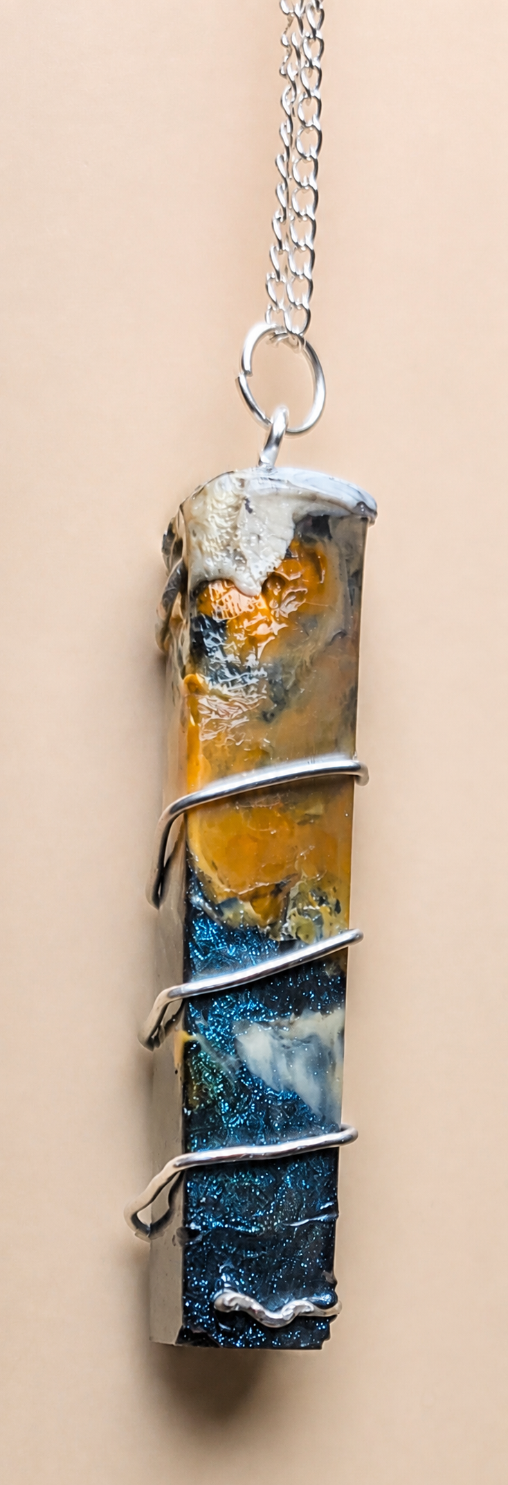 A vertical resin pendant necklace wrapped with silver wire, featuring a textured blue, white, and orange landscape design, hanging against a solid beige background.