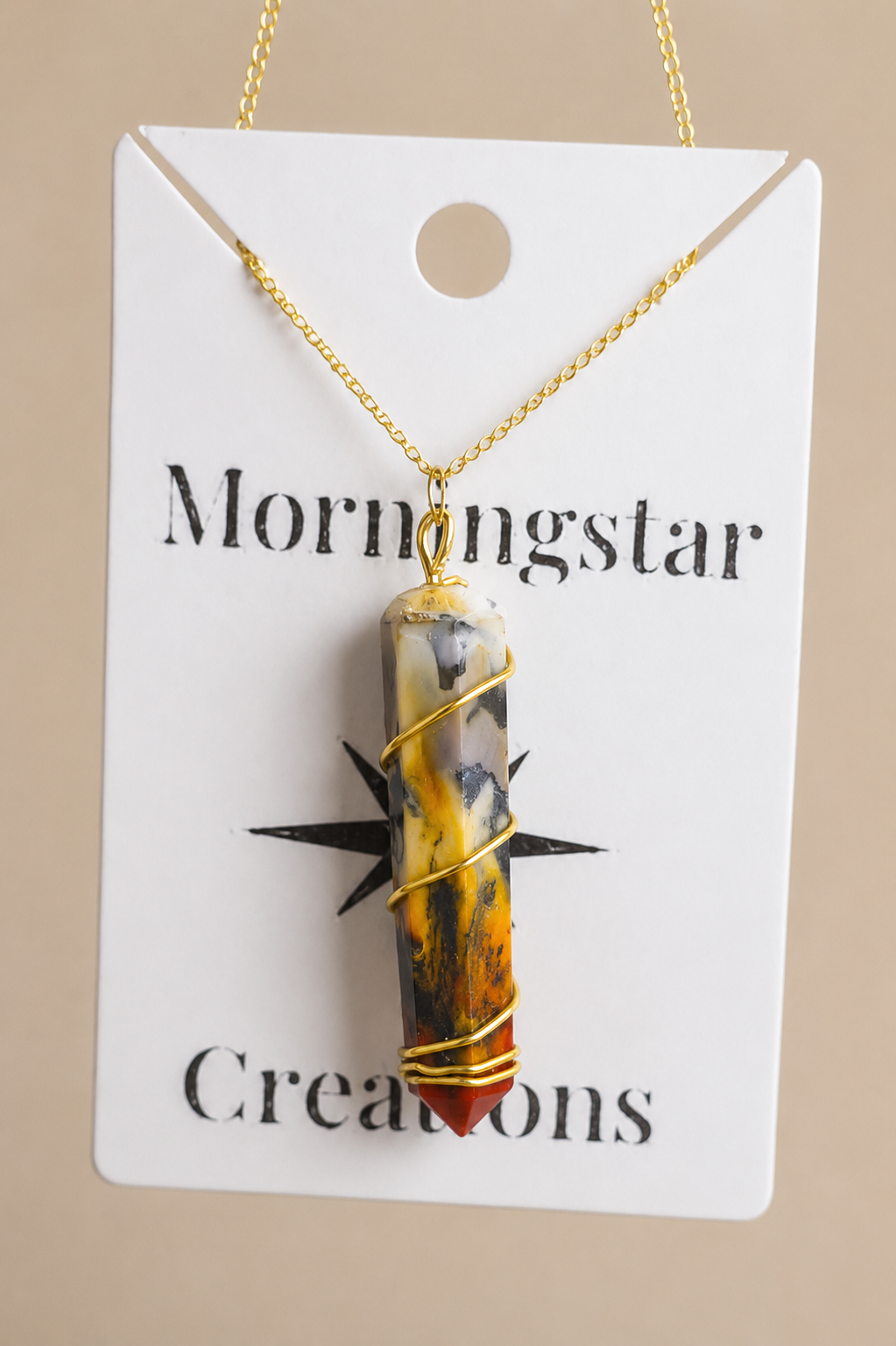 A gold chain necklace with a marbled amber, yellow, and gray stone pendant wrapped in gold wire, displayed on a white card labeled 'Mornigstar Creations' against a beige background.