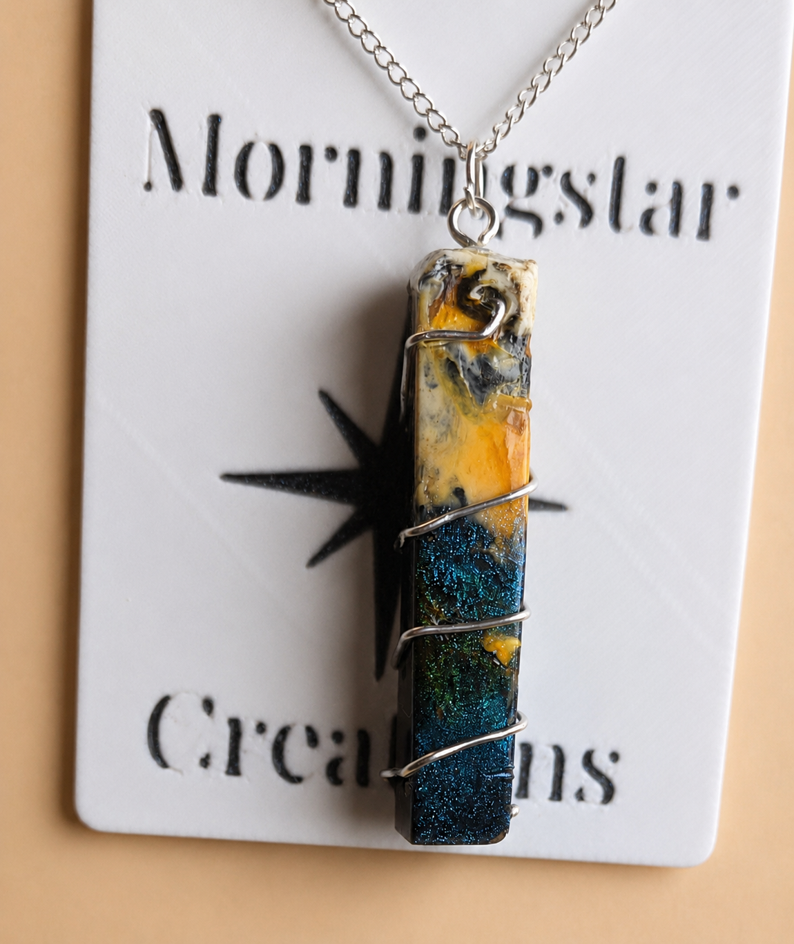 A rectangular wrapped pendant necklace with a marbled blue, yellow, and black design, suspended from a silver chain and displayed on a white card labeled 'Morningstar Creations'.
