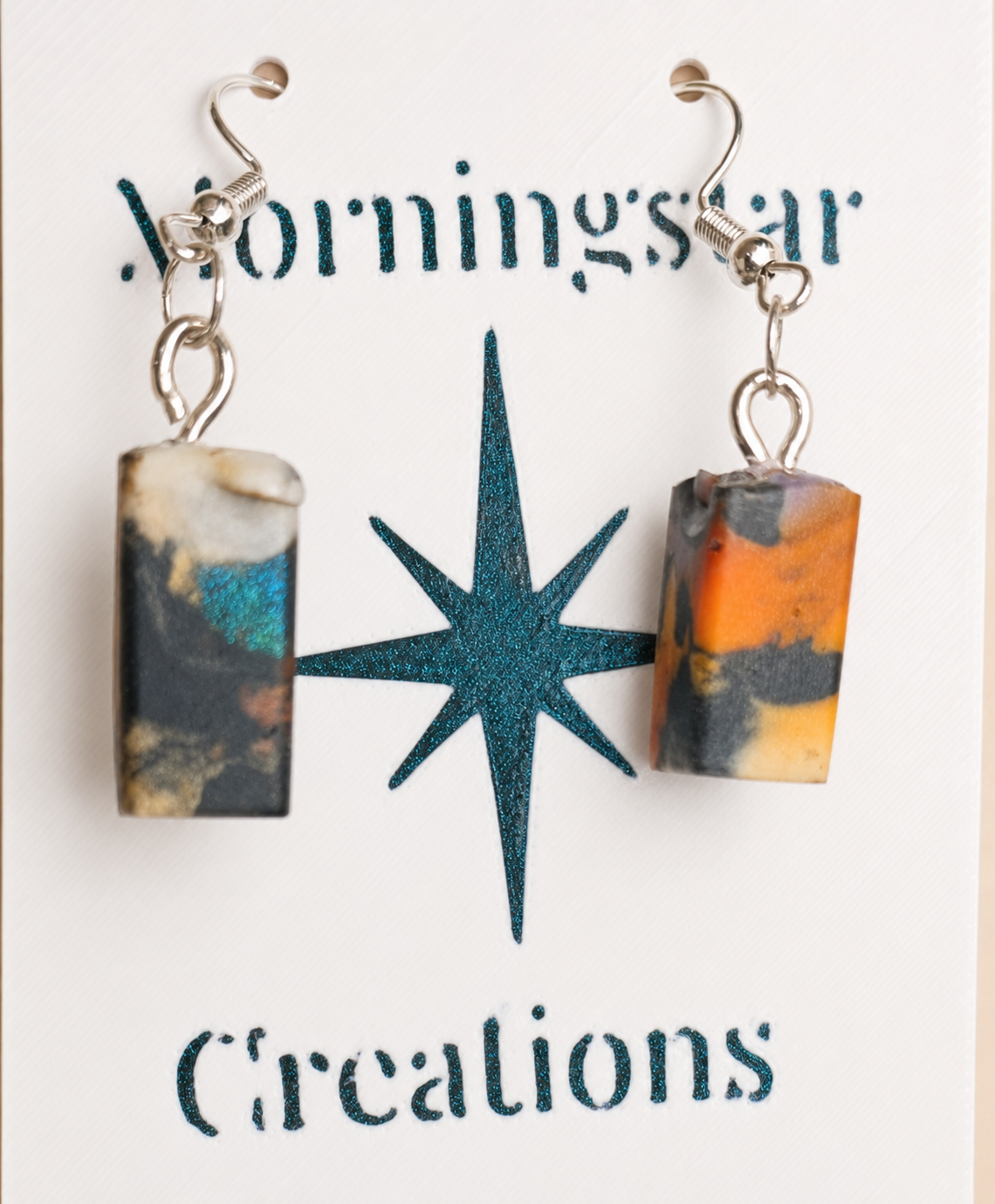 A pair of dangling earrings with rectangular marbled resin pendants in black, orange, and cream, hanging from silver fishhook wires on a white display card with the text 'MorningsTar Creations'.