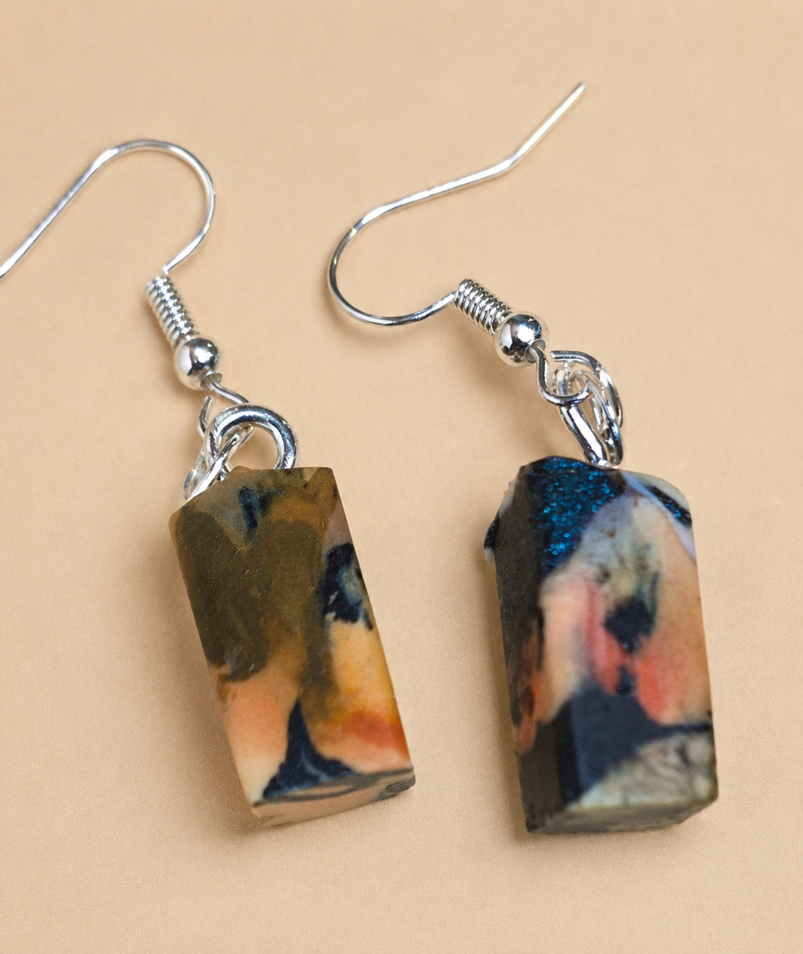 A pair of silver fishhook drop earrings with rectangular resin pendants featuring an abstract marbled pattern in black, gold, orange, and blue, set against a solid beige background.