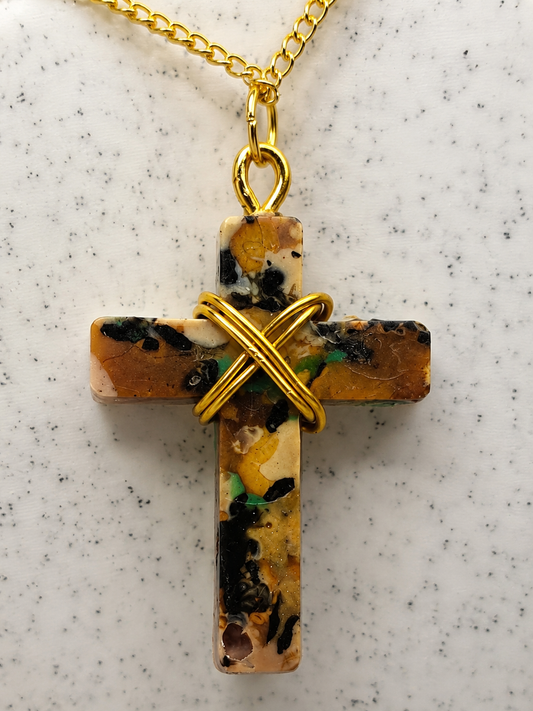 A cross-shaped pendant necklace made from a marbled stone with gold wire wrapping and a gold chain, displayed on a speckled white surface.