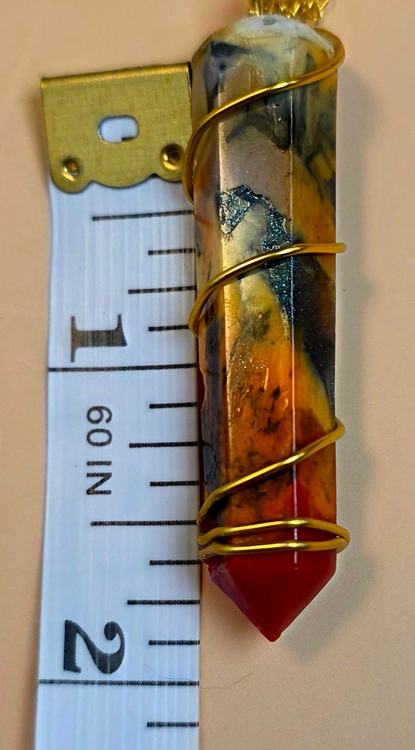 A wrapped crystal point with a red conical tip and marbled amber, gold, black, and blue-green body, shown next to a metal tape measure on a beige background.