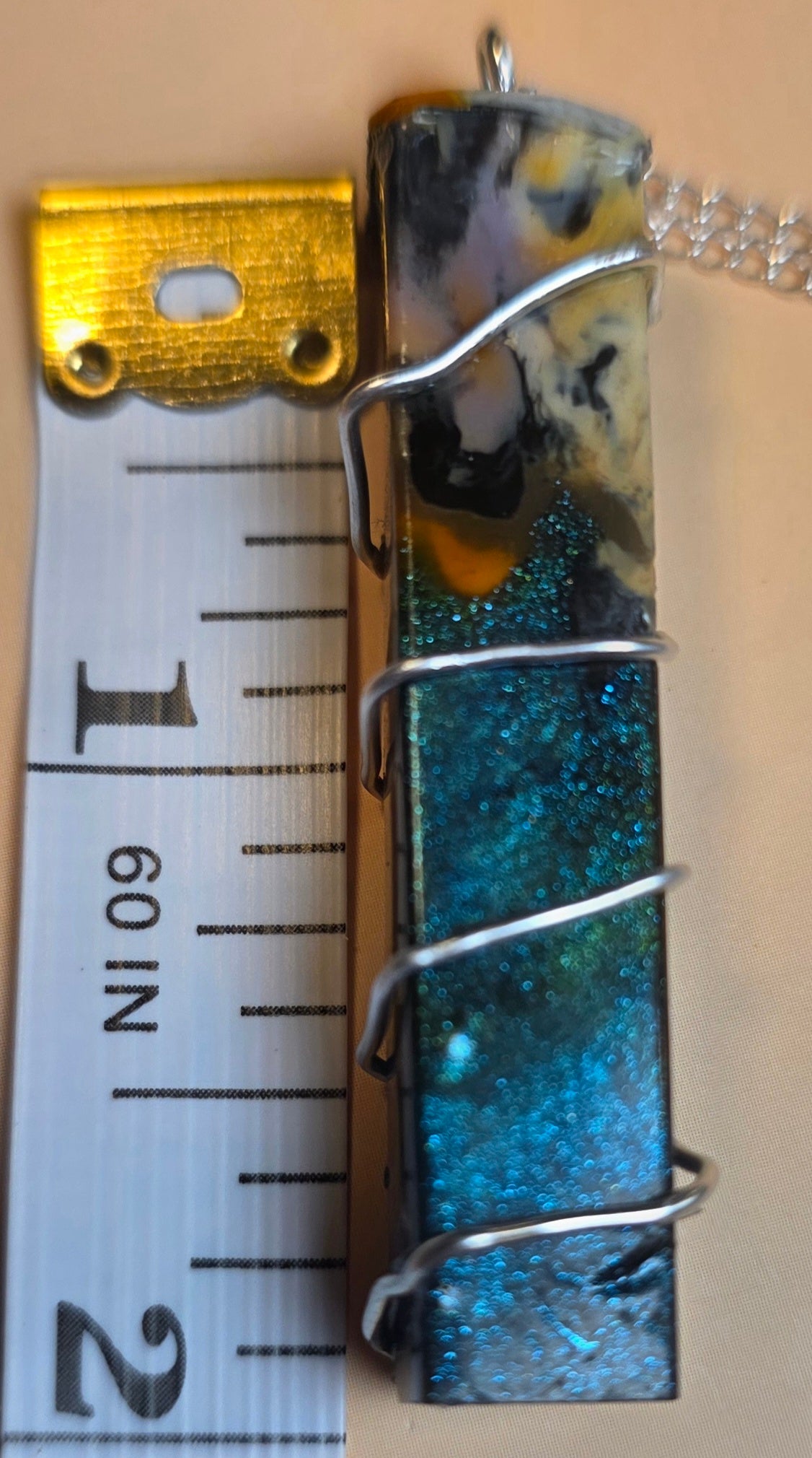 A rectangular resin pendant with swirling blue, teal, black, and yellow patterns, wrapped in silver wire with a gold cap and a silver chain, shown next to a ruler for scale.