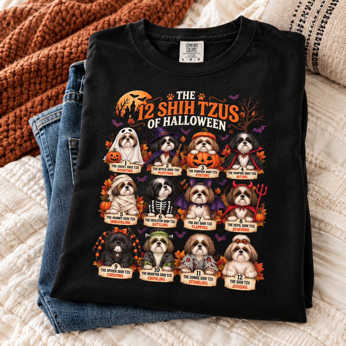 A folded black graphic T-shirt is displayed on top of blue jeans on a cream quilted bedspread, with a rust-orange knit blanket in the background. The shirt features a Halloween-themed graphic titled 'THE 12 SHIH TZUS OF HALLOWEEN' showing twelve Shih Tzu dogs dressed in various spooky costumes with names and descriptions below each.