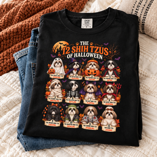 A folded black graphic T-shirt is displayed on top of blue jeans on a cream quilted bedspread, with a rust-orange knit blanket in the background. The shirt features a Halloween-themed graphic titled 'THE 12 SHIH TZUS OF HALLOWEEN' showing twelve Shih Tzu dogs dressed in various spooky costumes with names and descriptions below each.