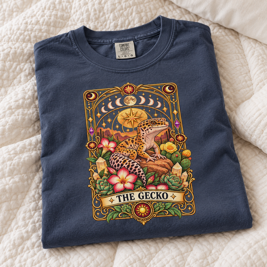 A navy blue crewneck t-shirt with a detailed 'The Gecko' tarot card graphic, laid flat on a cream-colored quilted blanket. The graphic features a leopard gecko, flowers, and celestial elements.