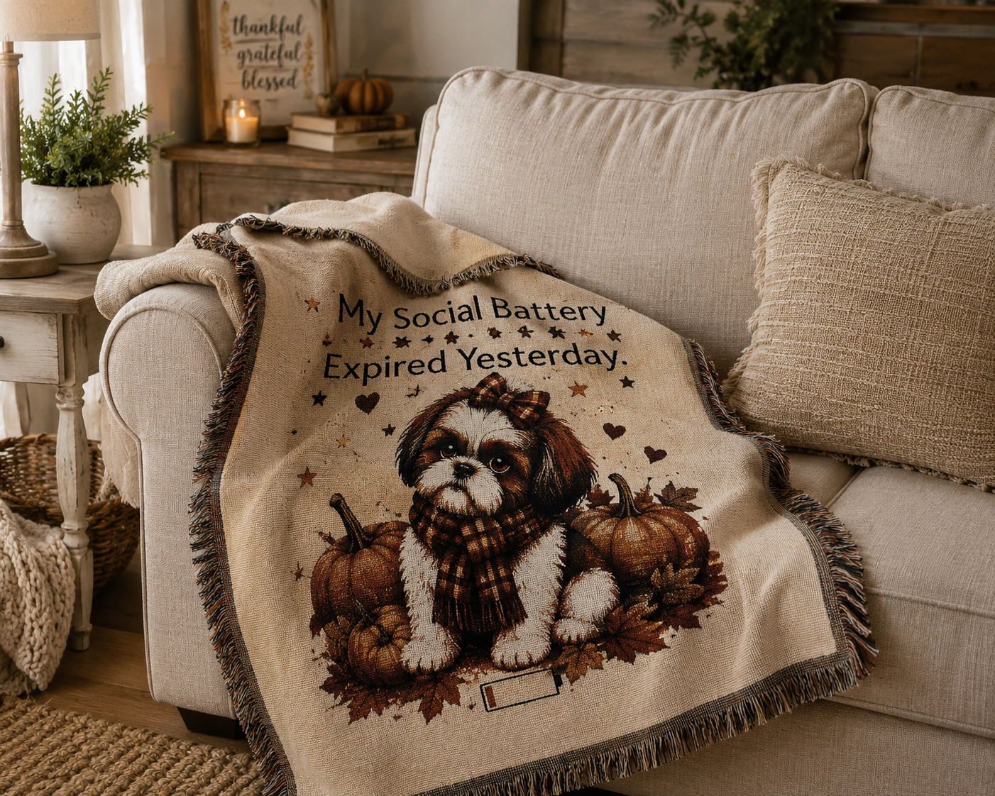 A cream throw blanket draped over a beige sofa in a cozy living room. The blanket features a printed illustration of a small Shih Tzu dog wearing a scarf, surrounded by pumpkins and autumn leaves, with the text 'My Social Battery Expired Yesterday.