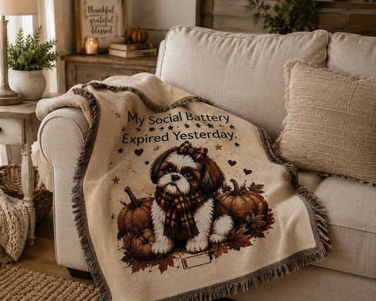 A cream throw blanket draped over a beige sofa in a cozy living room. The blanket features a printed illustration of a small Shih Tzu dog wearing a scarf, surrounded by pumpkins and autumn leaves, with the text 'My Social Battery Expired Yesterday.