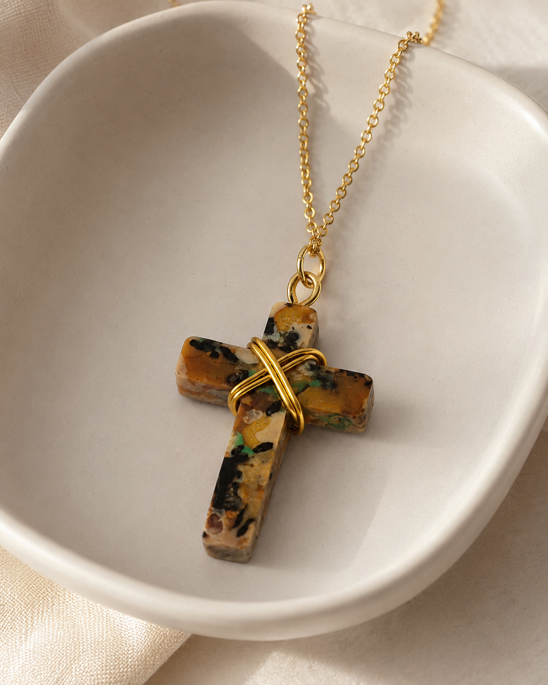 A gold chain necklace with a cross pendant made from a polished mottled stone in earthy brown, black, and green tones, wrapped with a gold wire accent, displayed inside a white ceramic bowl.