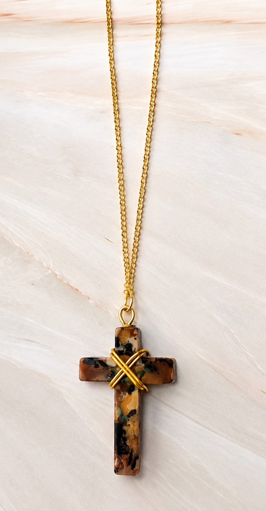 A gold chain necklace with a cross pendant made of brown and black marbled stone, accented with a gold wire cross on the front, displayed on a light beige marble background.