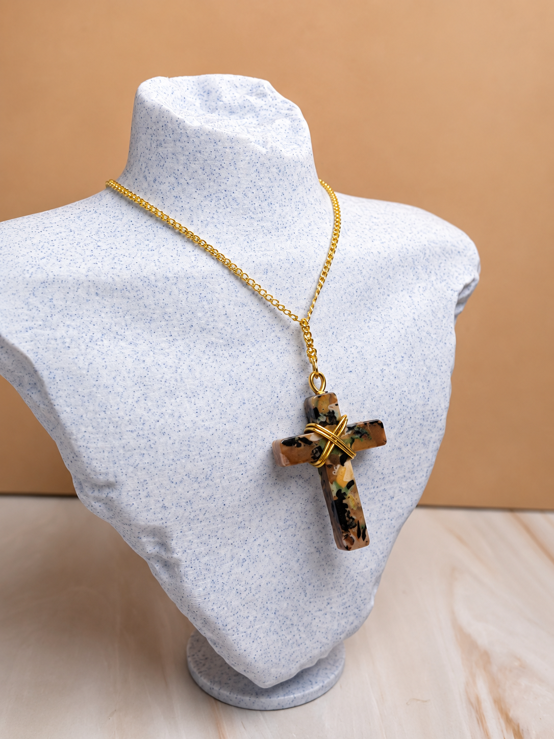A gold chain necklace with a cross pendant made of brown and black marbled stone, wrapped with a gold wire accent at the center, displayed on a light gray fabric mannequin bust against a warm beige background.