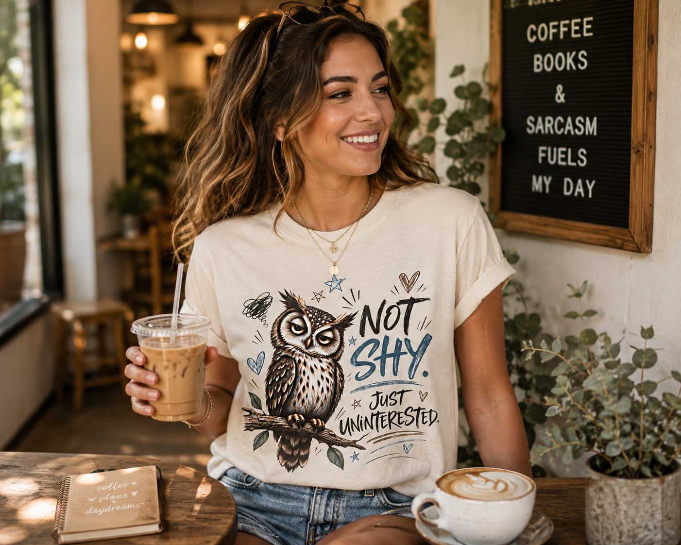 A woman sitting at a cafe table wearing a cream-colored graphic t-shirt with an owl illustration and the text 'NOT SHY. JUST UNINTERESTED.