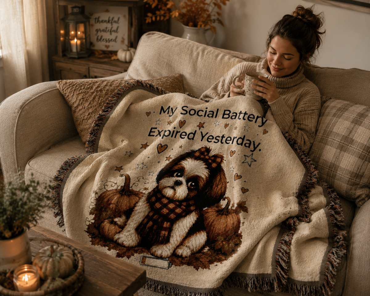 A cozy living room scene with a woman reclining on a beige couch wrapped in a cream throw blanket featuring a Shih Tzu dog illustration and the text 'My Social Battery Expired Yesterday.' The blanket has fringe edges and is styled with autumn decor, candles, and plaid pillows.