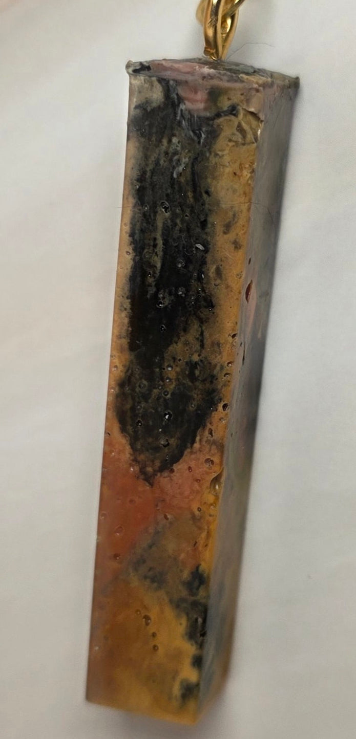 A rectangular bar pendant with a gold chain, featuring a marbled pattern of black, brown, and gold, resting on a light off-white background.