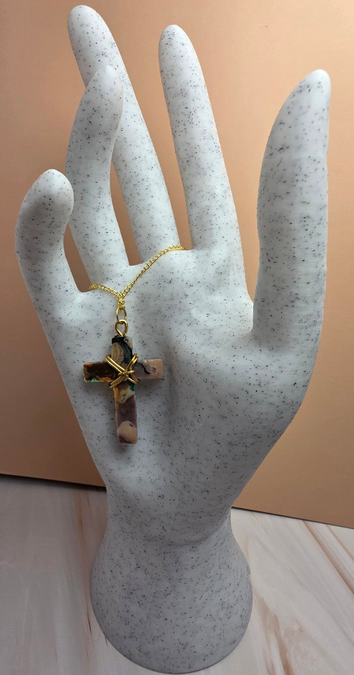 A gold cross pendant with a pink and green stone inlay, displayed on a gray speckled hand mannequin against a peach-colored background.
