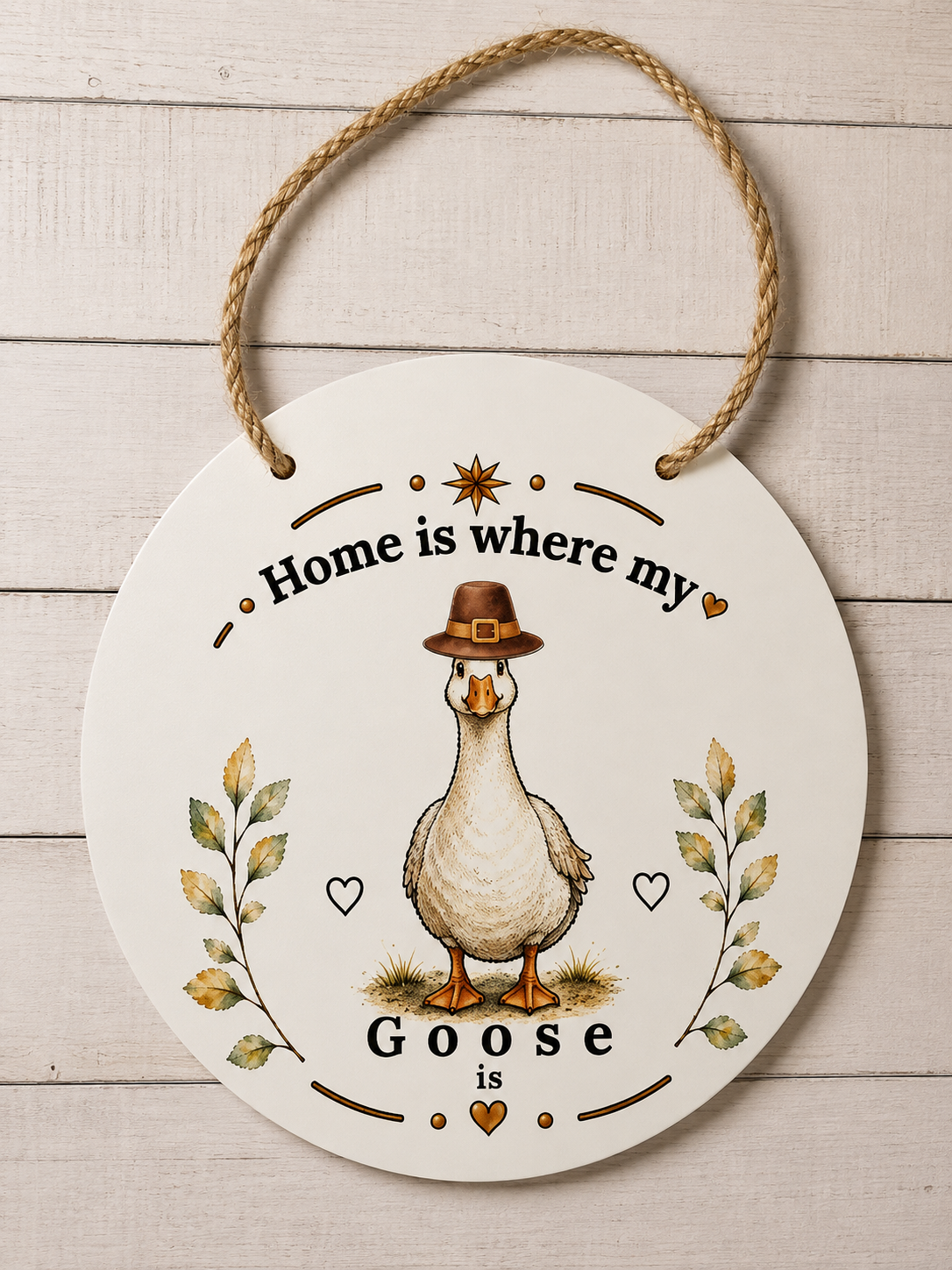 Round white hanging wall sign with a rope loop, featuring a cartoon goose wearing a brown hat and the text 'Home is where my Goose is' on a whitewashed wood plank background.