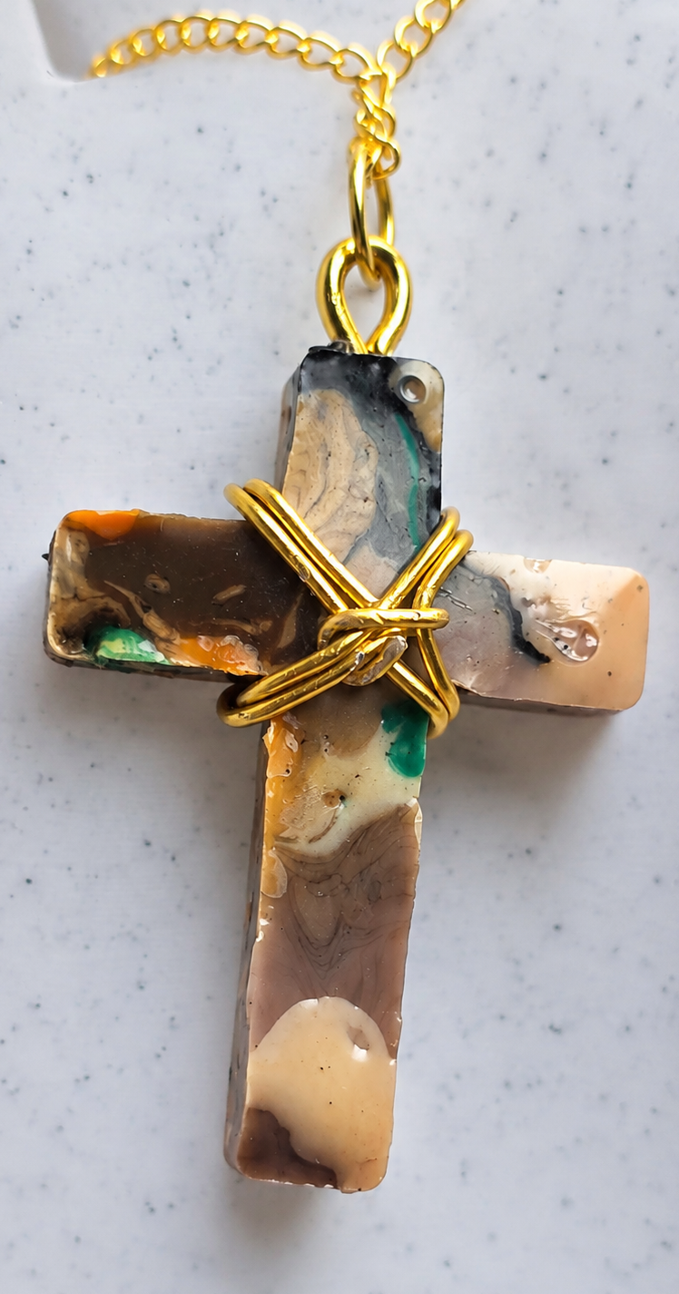 A handmade cross pendant necklace made from layered marbled resin with brown, beige, black, and green tones, wrapped with a gold wire cross and hanging from a gold chain on a speckled white surface.