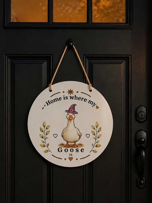 A round white Halloween wall hanging sign with a goose illustration and the phrase “Home is where my Goose” hangs on a black front door. The scene is decorated for Halloween with carved pumpkins, a witch hat, a “Welcome to Witches” doormat, and warm lantern light.
