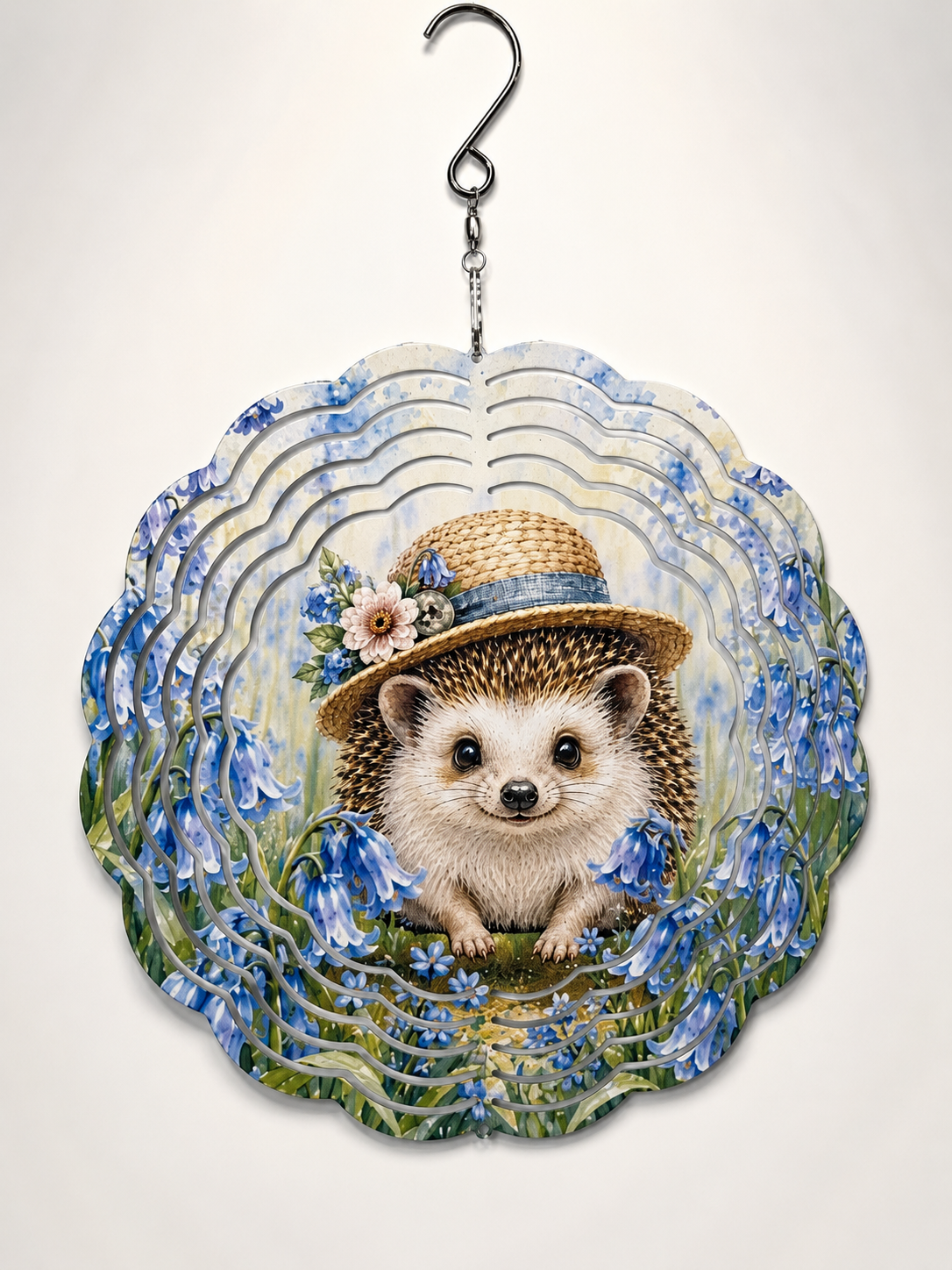 A scalloped decorative wind spinner featuring an illustrated hedgehog wearing a straw hat with flowers, surrounded by blue bellflowers and green foliage, suspended from a metal hook against a plain light background.