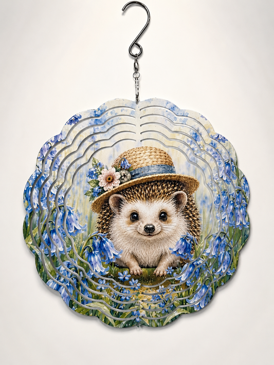 A scalloped decorative wind spinner featuring an illustrated hedgehog wearing a straw hat with flowers, surrounded by blue bellflowers and green foliage, suspended from a metal hook against a plain light background.