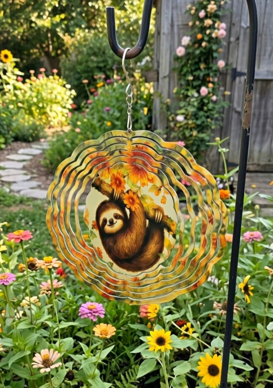 A circular metal garden wind spinner featuring a sloth illustration and autumn leaves, hanging on a black metal stake in a colorful garden.