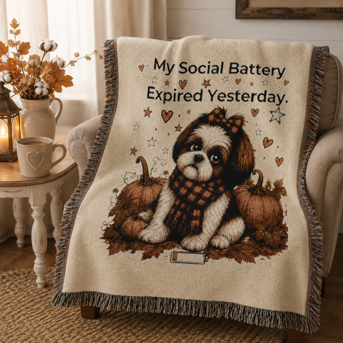 A cozy woven throw blanket draped over a beige armchair in a warm living room. The blanket features a Shih Tzu dog wearing a plaid scarf, surrounded by pumpkins, with the text 'My Social Battery Expired Yesterday.