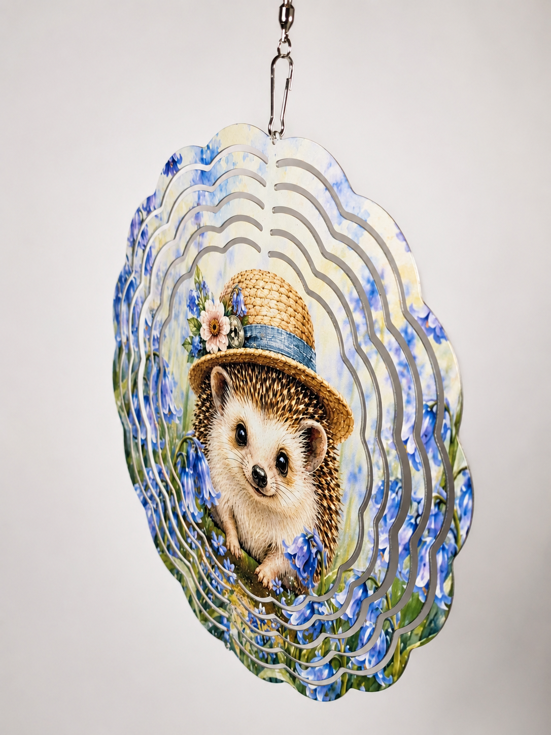 A decorative hanging wind spinner with a scalloped edge, featuring a printed illustration of a hedgehog wearing a straw hat surrounded by blue flowers, with layered wavy cutout patterns in the background.