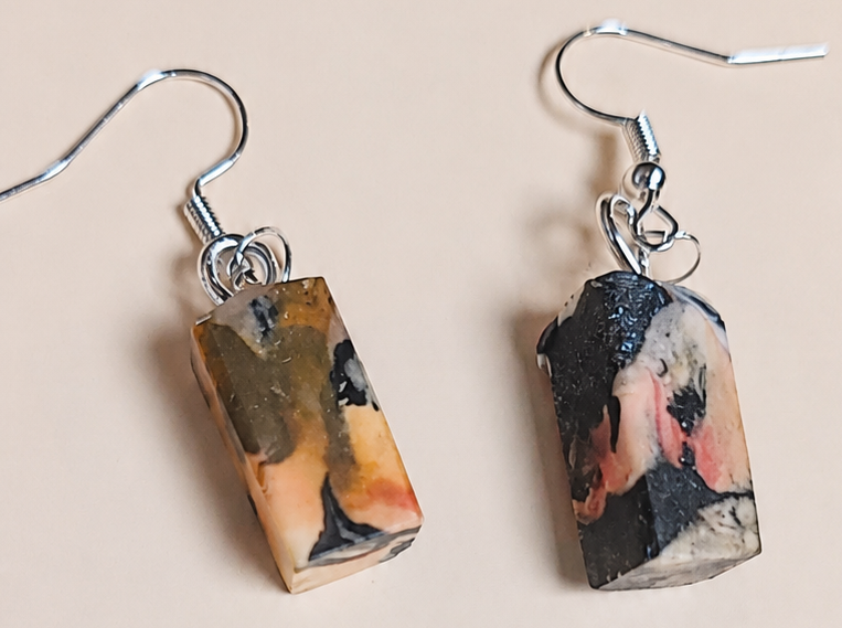 A pair of silver fishhook earrings with rectangular resin pendants featuring a painted portrait design in black, pink, and gold tones, displayed on a solid light beige background.
