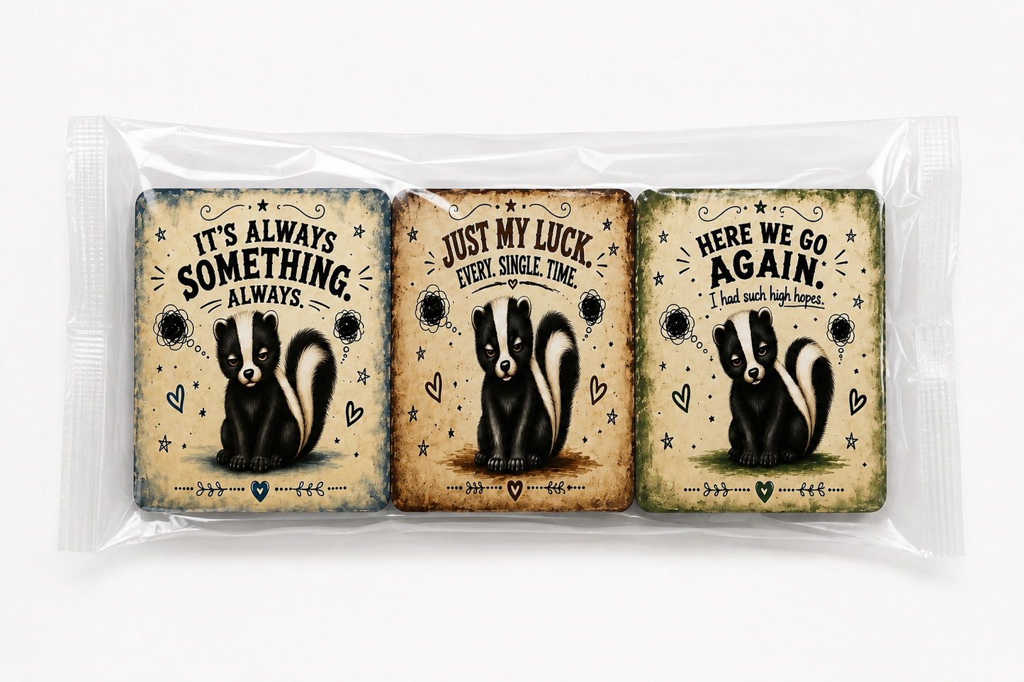 Three rectangular printed cards or stickers in a clear plastic sleeve, each featuring a skunk illustration and a humorous quote: 'IT'S ALWAYS SOMETHING ALWAYS,' 'JUST MY LUCK EVERY SINGLE TIME,' and 'HERE WE GO AGAIN I had such high hopes.'.