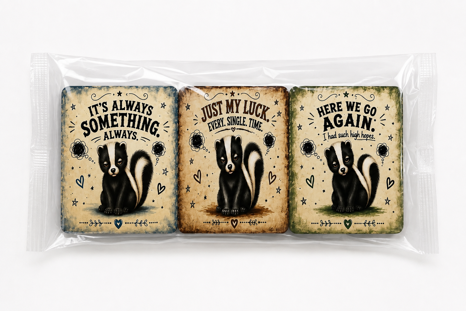 Three rectangular printed cards or stickers in a clear plastic sleeve, each featuring a skunk illustration and a humorous quote: 'IT'S ALWAYS SOMETHING ALWAYS,' 'JUST MY LUCK EVERY SINGLE TIME,' and 'HERE WE GO AGAIN I had such high hopes.'.