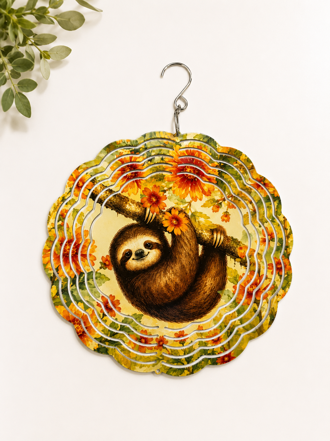 A scalloped circular hanging sign featuring a sloth hanging from a branch with sunflowers, set against a floral pattern. The sign has a silver metal hook at the top and is displayed against a clean white background with a small green plant leaf in the upper left corner.