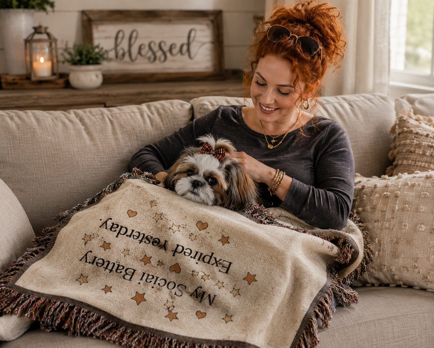 A woman with red hair sits on a beige couch holding a small dog under a cream throw blanket with embroidered text and star motifs. The blanket reads 'My Social Battery Expired Yesterday.