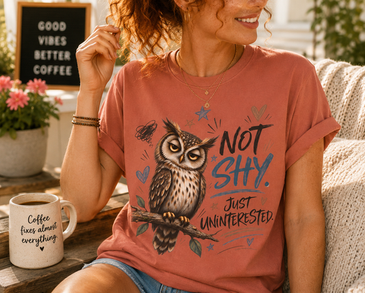 A woman wearing a terracotta-colored graphic t-shirt with an owl illustration and the text 'NOT SHY. JUST UNINTERESTED.