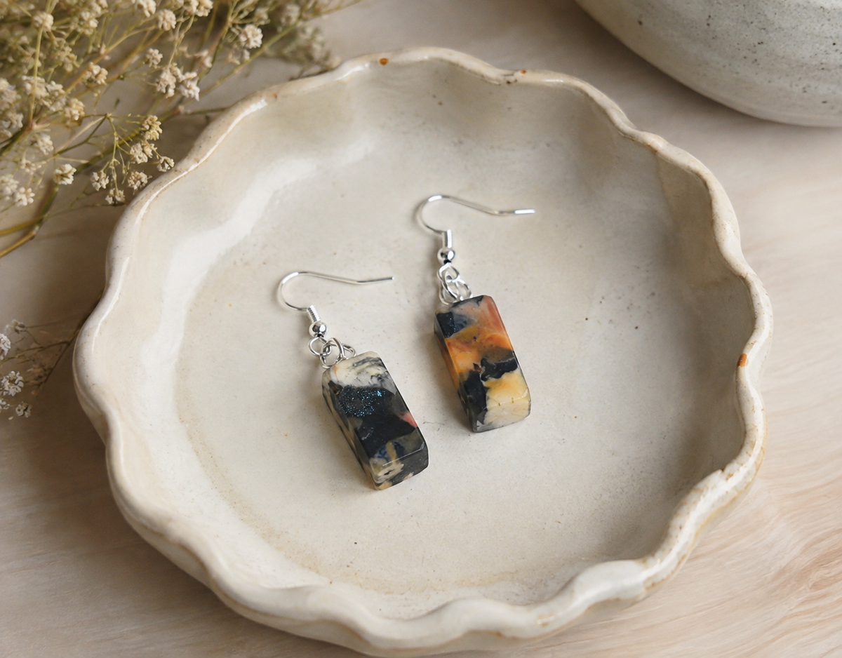 A pair of rectangular dangle earrings with marbled black, orange, and cream resin pendants on silver fishhook wires, displayed on a scalloped ceramic dish.
