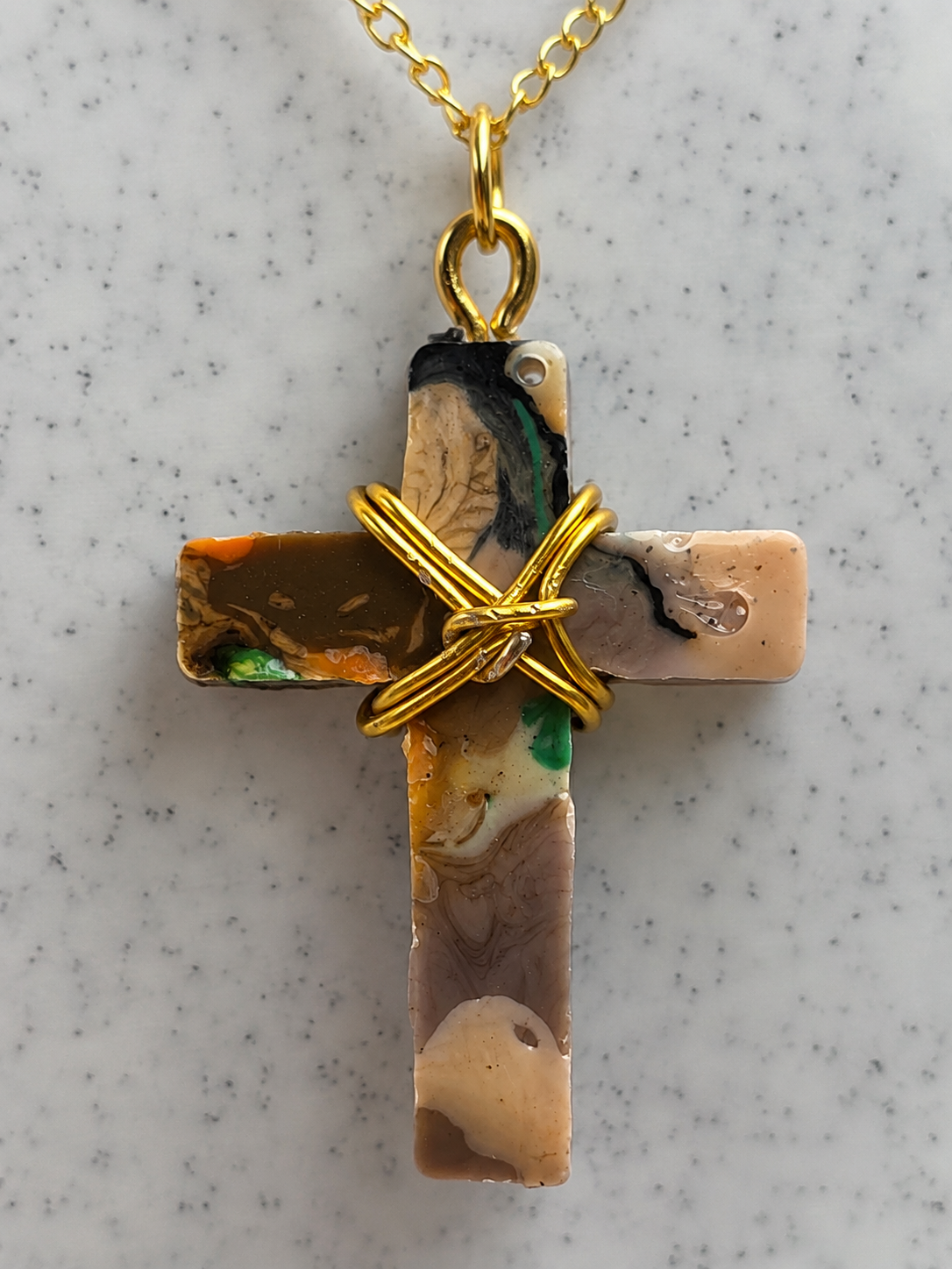 A gold cross pendant necklace featuring a marbled stone inlay with beige, brown, black, and green swirls, wrapped in a gold wire cross at the center, hanging from a gold chain on a speckled white marble background.